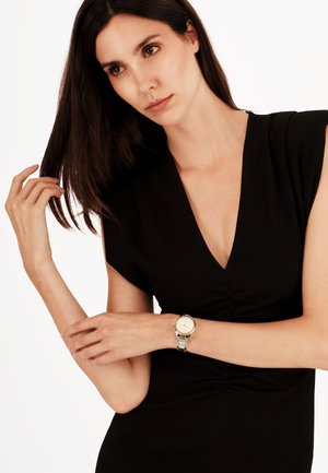 Sort kjole med dyb V-udskæring og korteærmer, med subtil rynkning ved taljen. Et sølvur med rund urskive pryder håndleddet.