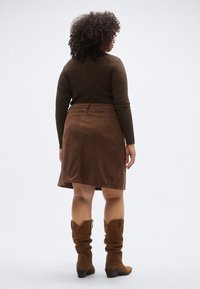 Fustă maro din catifea cu talie strâmtă, asortată cu un top cu mâneci lungi și imprimeu și cu cizme maro de cowboy, largi.