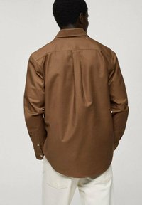 Brown button-up shirt with a smooth texture, back pleat detail, and a classic collar, paired with light-colored pants.