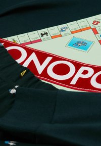 Tela temática de Monopoly en verde con un parche bordado en rojo que dice "MONOPOLY", tela oscura con manga acampanada y pequeños personajes de dibujos animados.
