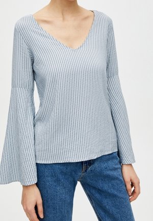 Femme portant un haut à manches longues bleu clair à rayures verticales blanches avec des manches évasées et un jean bleu.