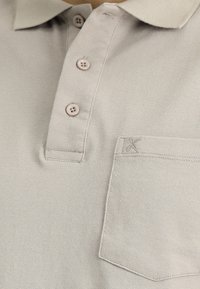 Polo shirt beige chiaro con tessuto texturizzato, tre bottoni e una piccola tasca con dettaglio ricamato.