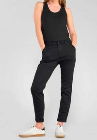 Femme portant un haut noir sans manches, un pantalon noir taille haute et des baskets blanches, debout les mains dans les poches.