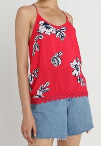 Top de tirantes espagueti de color rojo con estampados florales en blanco y azul, con un borde de encaje en la parte inferior, combinado con shorts de mezclilla.