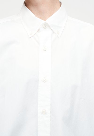 Άνδρας που φοράει απλό λευκό πουκάμισο με κουμπιά και γιακά με κουμπιά, ορατός από το λαιμό μέχρι το μέσο του κορμού, μπροστά σε λευκό φόντο.