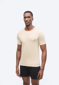 Mand stående iført en beige T-shirt med korte ærmer med "BOSS"-logo og sorte shorts foran en ensfarvet lysegrå baggrund.