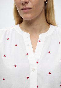 Γυναίκα που φοράει λευκό πουκάμισο με μικρές κόκκινες κεντημένες καρδιές, στρογγυλή λαιμόκοψη με οπίσθια σχισμή και ένα μόνο περλέ κουμπί, καθώς και ασημένια σκουλαρίκια.