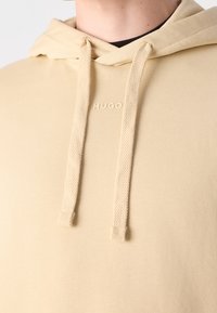 Sudadera beige de tela suave, con un bolsillo frontal tipo canguro, cordones ajustables y un pequeño logo en relieve en el pecho.