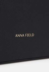 Surface texturée noire avec le texte en relief doré "ANNA FIELD" centré près du bord inférieur.