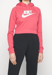 Růžová krátká mikina s kapucí, dlouhými rukávy a bílým logem Nike na přední straně. Kombinováno s tmavě zbarvenými tepláky. Hladká textura.