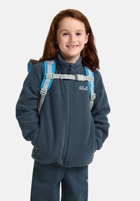 Chaqueta de forro polar gris oscuro con cuello alto, que presenta una cremallera frontal y herrajes grises. Se ven correas de mochila en azul claro.
