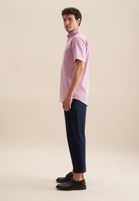 Camicia rosa a maniche corte con bottoni e tasca sul petto, abbinata a pantaloni navy scuri e scarpe nere, mostrata in un profilo verticale.