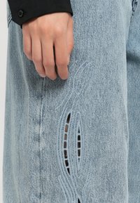 Blugi din denim de un albastru deschis, cu broderii și decupaje în formă de val pe coapsă, asortați cu o cămașă neagră cu nasturi.