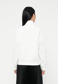 Donna con lunghi capelli neri che indossa un pullover bianco a collo alto e una gonna nera plissettata, vista di spalle contro uno sfondo bianco uniforme.