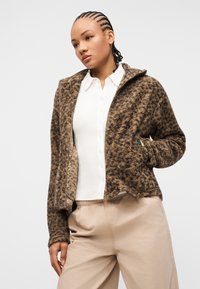 Γυναίκα με πλεξούδες, ντυμένη με καφέ ανάγλυφο μπουφάν με φερμουάρ πάνω από λευκό πουκάμισο με γιακά και μπεζ παντελόνι, στέκεται μπροστά σε απλό φόντο.