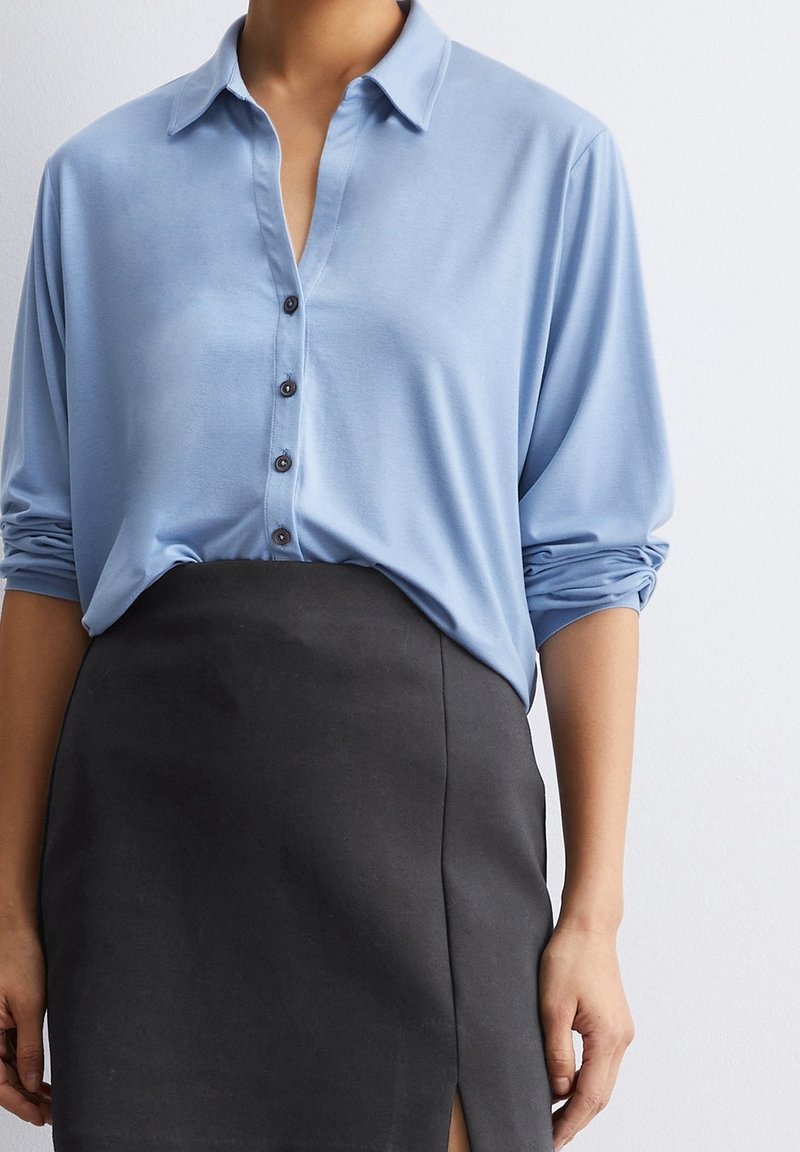 Personne portant une chemise bleu clair à boutons avec les manches retroussées et une jupe noire taille haute avec une fente sur le devant.