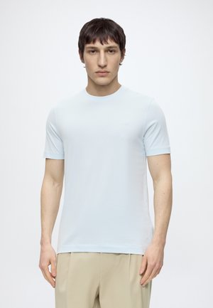 Jeune homme aux cheveux courts et foncés portant un t-shirt uni bleu clair à manches courtes et un pantalon beige, debout devant un fond blanc.