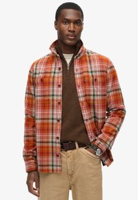 Camisa de franela a cuadros en tonos naranjas, rojos y verdes, con botones en el frente, dos bolsillos en el pecho y un cuello mandarín. Usada sobre un suéter marrón.