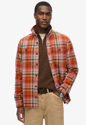 Camisa de franela a cuadros en tonos naranjas, rojos y verdes, con botones en el frente, dos bolsillos en el pecho y un cuello mandarín. Usada sobre un suéter marrón.