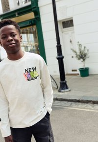 Lysegrå langærmet sweatshirt med farverigt "NEW YORK"-motiv, båret med mørke bukser, sat op mod en gadebaggrund.
