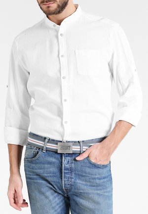 Mann trägt ein weißes Hemd mit hochgekrempelten Ärmeln und blaue Jeans mit einem gestreiften Levi's-Gürtel, Hand in der Hosentasche.