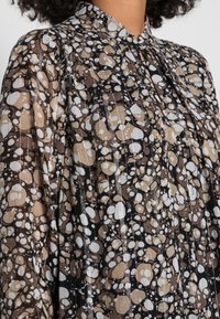 Genomskinlig brun blus med ett svart, krämfärgat och beige abstrakt mönster. Har krage och vertikala nålrandiga ränder på tyget.