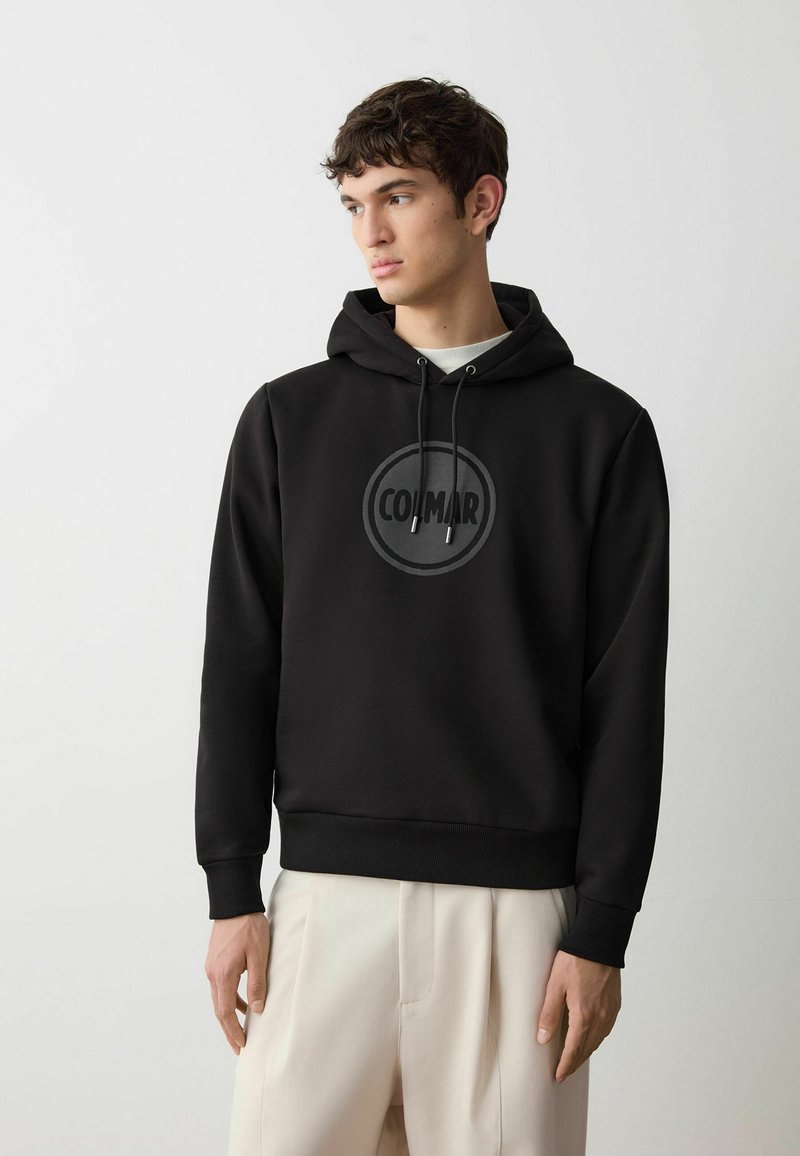 Felpa nera con cappuccio e logo circolare frontale. Realizzata in tessuto liscio, presenta lacci e polsini a coste. Indossata con pantaloni chiari.