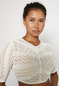 Top court en dentelle blanche avec des motifs ondulés texturés, manches courtes et design ajusté, présentant un ourlet à volants et une légère brillance.