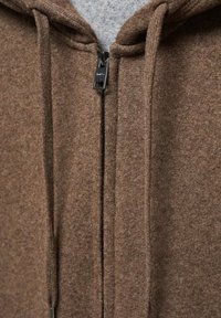 Sweat à capuche zippé marron en tissu doux et texturé. Comprend une fermeture éclair métallique et des cordons de serrage. Capuche doublée d'un matériau gris plus clair.