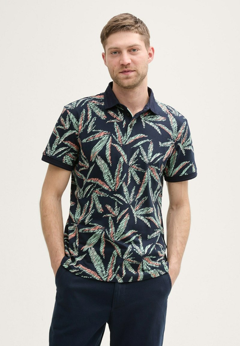 Marineblauwe poloshirt met een bladerenpatroon in groen en rood. Heeft een effen kleur kraag en korte mouwen. Gemaakt van een gladde stof.