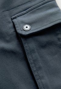 Dunkelgrauer Stoff mit glatter Textur, ausgestattet mit einer kontrastierenden Klappentasche und einem Metall-Druckknopf mit geprägtem Branding.