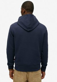 Marineblauwe hoodie met een grote verstelbare capuchon, geribbelde manchetten en zoom. Heeft een gladde textuur en een ontspannen pasvorm, zichtbaar vanaf de achterkant.