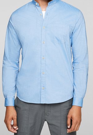 Mann trägt ein hellblaues, langärmliges Hemd mit Stehkragen und graue Hose, steht vor einem schlichten Hintergrund.