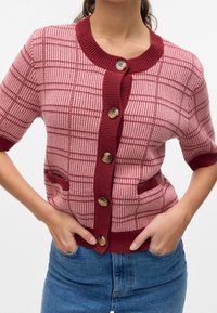 Femme portant un cardigan en tricot à manches courtes à carreaux roses et bordeaux avec boutons et poches avant, associé à un jean bleu.