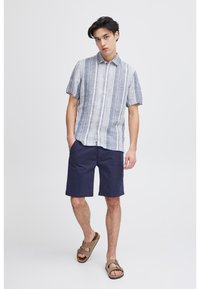 Chemise légère à rayures bleues et blanches à manches courtes, associée à un short marine. Le mannequin porte des sandales en cuir marron. Design minimaliste.