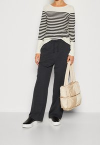 Person klädd i en svart och vit randig långärmad topp, mörka vida byxor, svarta och vita platåskor, som håller en beige quiltad toalettväska.
