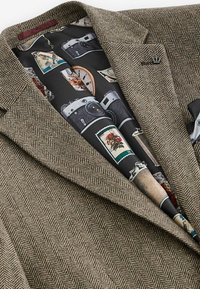 Blazer marron en chevron avec doublure noire comportant des illustrations d’appareils photo vintage et de timbres-poste.