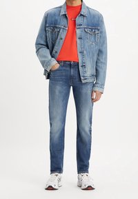 Chaqueta de mezclilla en azul claro con dos bolsillos en el pecho, sobre una camiseta de cuello redondo roja, complementada con jeans azules y zapatillas blancas.