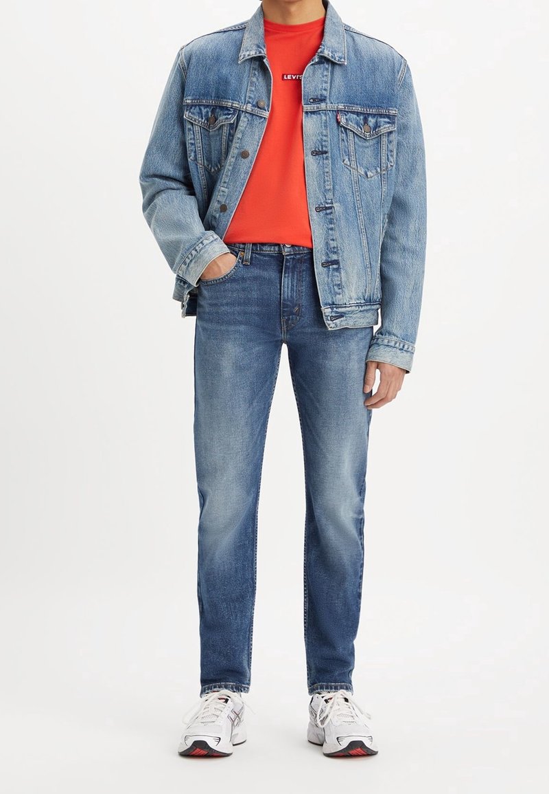Chaqueta de mezclilla en azul claro con dos bolsillos en el pecho, sobre una camiseta de cuello redondo roja, complementada con jeans azules y zapatillas blancas.