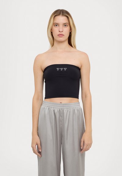 Crop top noir sans bretelles avec un design ajusté, arborant les chiffres blancs imprimés "777" à l'avant, porté avec un pantalon gris ample.