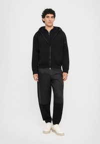 Svart hettegenser med glidelås og hette, kombinert med svarte bukser med en glinsende tekstur og to-farget design, samt hvite snøresko.
