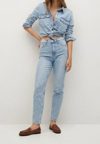 Camisa de mezclilla de color azul claro atada a la cintura, con dos bolsillos en el pecho. Combinada con jeans de tiro alto en azul claro y mocasines marrones sin talón.