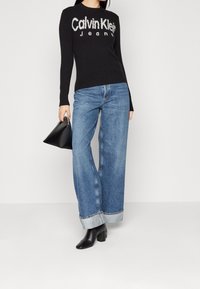 Pull en maille noire avec logo "Calvin Klein Jeans", associé à un jean large bleu clair et des bottines noires à talons. Sac à main noir simple.