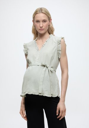 Gravid kvinde iført en lysegrønternet ærmeløs bluse med flæser og sorte bukser, stående foran en ensfarvet baggrund.