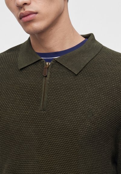 Gros plan d'une personne portant un pull zippé texturé vert foncé avec un col sur une chemise rayée bleue et blanche.