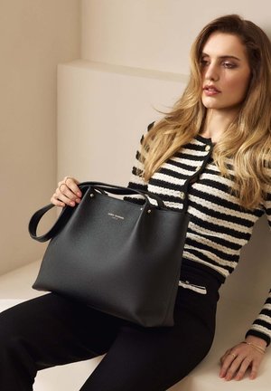 Sac fourre-tout en cuir noir avec une texture grainée, doté de poignées courtes et de subtils accents dorés. Le modèle porte un pull rayé.
