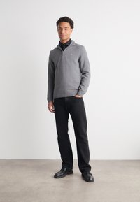 Grau zip-fester Pullover mit geripptem Saum, getragen über einem Hemd mit Kragen, kombiniert mit schwarzen Hosen und schwarzen Schuhen, auf einem neutralen Hintergrund stehend.
