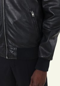 Zwarte leren bomberjack met een gladde textuur, een ritszak, ribgebreide manchetten en kraag. Voorzien van een metallic ritssluiting en een getailleerd ontwerp.
