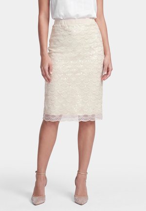 Donna che indossa una gonna beige con paillettes al ginocchio con orlo in pizzo smerlato, abbinata a una camicetta bianca e tacchi alti beige con cinturino alla caviglia.