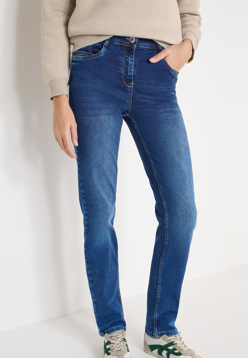 Jean bleu denim à coupe skinny avec un design délavé, comprenant cinq poches, une fermeture éclair et un bouton. Associé à des baskets vertes et beiges.